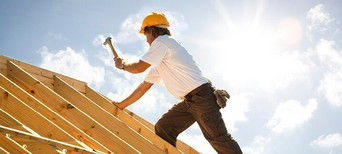 Dachdecker mit gelbem Schutzhelm beim Befestigen von Konstruktionsvollholz (KVH) auf einem Dachstuhl unter strahlendem Himmel.